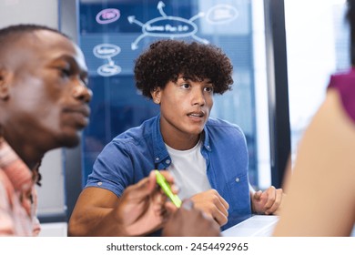 A young biracial man with curly brown hair is discussing with colleagues in a modern business office. He's wearing a blue shirt, holding a green marker, and has a thoughtful look, unaltered. - Powered by Shutterstock