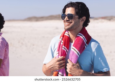 Young biracial man enjoys a sunny beach day, with copy space. He outdoors, smiling with sunglasses and a towel around his neck. - Powered by Shutterstock