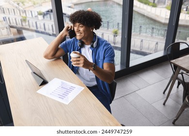 A young biracial man holding coffee, talking on the phone, sitting by a laptop in a modern business office. He has curly dark hair, wearing a blue shirt, enjoying a break, unaltered. - Powered by Shutterstock