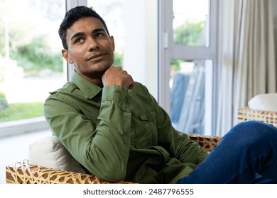 Young biracial man sits thoughtfully in a home setting, unaltered. He appears contemplative, relaxing on a wicker chair near a bright window. - Powered by Shutterstock