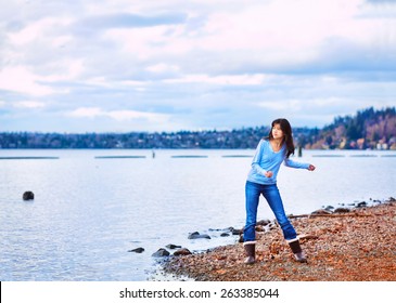 Young biracial teen girl in blue shirt and jeans along rocky lake shore, throwing rocks into the water on cloudy overcast day - Powered by Shutterstock