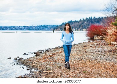 Young biracial teen girl in blue shirt and jeans walking along rocky shoreline of lake in early spring or fall - Powered by Shutterstock