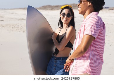 Young biracial woman and African American man enjoy a sunny beach day. They carry a surfboard, ready for an exciting surfing session outdoors. - Powered by Shutterstock