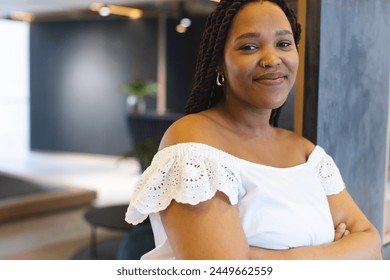 A young biracial woman with braided hair is sitting, arms crossed, in a modern business office. She wears a white off-shoulder top and is smiling gently, unaltered. - Powered by Shutterstock