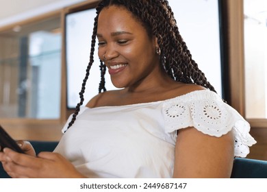 A young biracial woman with braided hair smiling at her phone. She wearing white off-shoulder top and sitting in a well-lit room in a modern business office, unaltered - Powered by Shutterstock