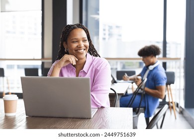 Young biracial woman with braided hair working on laptop, smiling, in a modern business office. She wears a pink shirt; coffee cup on table, man in background, unaltered. - Powered by Shutterstock