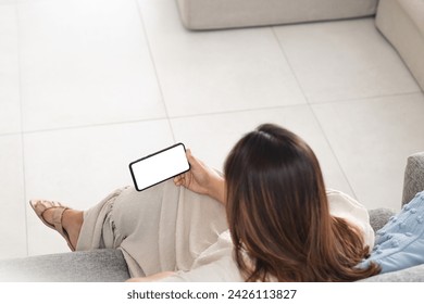 Young biracial woman relaxes on a couch at home, holding a smartphone with a blank screen. She's enjoying a casual day indoors, browsing or texting. - Powered by Shutterstock