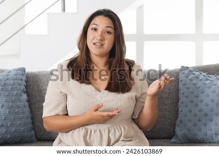 Young biracial woman sits comfortably at home on a couch on a video call. She gestures while speaking, adding emphasis to her conversation.