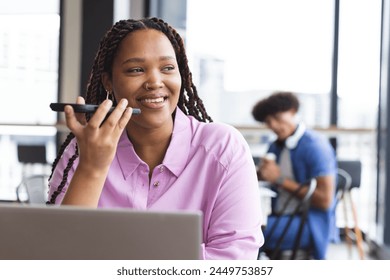 Young biracial woman talking on smartphone, working on laptop in a modern business office. She has braided hair, wears a pink shirt, and a young biracial man is in the background, unaltered. - Powered by Shutterstock