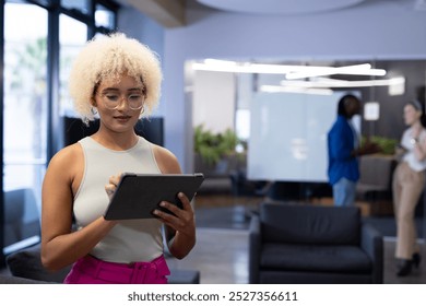 Young biracial woman using tablet at office as colleagues chat in the background, copy space. Bright, open workspace with large windows and contemporary furniture, creating modern feel, unaltered - Powered by Shutterstock