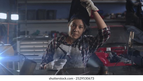 A young biracial woman is working in workshop, holding a welding tool. She has light brown skin, dark hair tied up, and is wearing a protective apron - Powered by Shutterstock