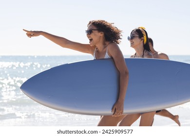 Young biracial women carry a surfboard on the beach. Excitement fills the air as they prepare for a day of surfing. - Powered by Shutterstock