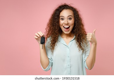 Young black african american fun surprised satisfied happy curly driver woman 20s wearing blue shirt holding car key show thumb up like gesture isolated on pastel pink color background studio portrait