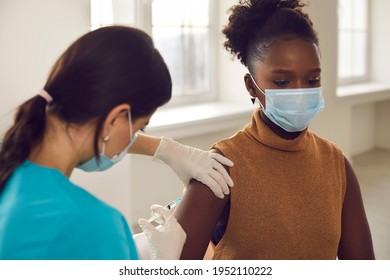 Young Black African American Woman Patient In Protective Face Mask Sitting Getting Vaccination Injection Against Coronavirus From Female Nurse In Clinic. Vaccinating Against COVID-19 Infection