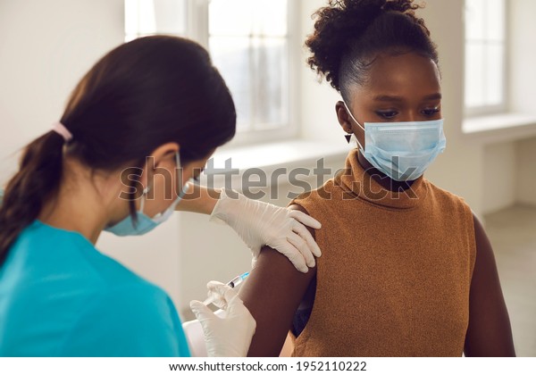 Young black african american woman patient in\
protective face mask sitting getting vaccination injection against\
coronavirus from female nurse in clinic. Vaccinating against\
COVID-19 infection