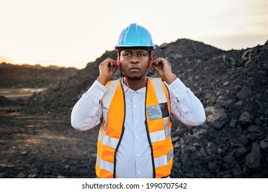 A young Black African coal mine foreman looking into camera wearing reflective bib and hard hat is putting in ear protection after a long day of work on site at the coal mine - Powered by Shutterstock