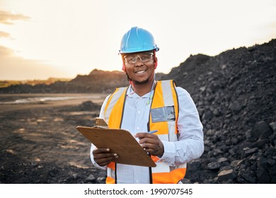 A young Black African coal mine foreman looking into camera smiling wearing reflective bib and hard hat is inspecting samples of coal on his clipboard after a long day of work on site at the coal mine - Shutterstock ID 1990707545