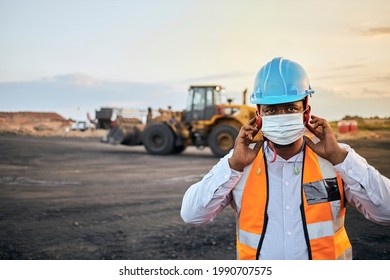 A Young Black African Coal Mine Foreman Wearing Reflective Bib And Hard Hat Is Putting On A Covid 19 Face Mask After A Long Day Of Work On Site At The Coal Mine