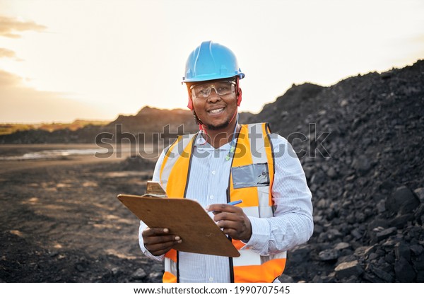 A young Black African coal mine foreman looking into\
camera smiling wearing reflective bib and hard hat is inspecting\
samples of coal on his clipboard after a long day of work on site\
at the coal mine