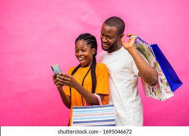 Young black African couple holding shopping bags taking selfies standing on a pink studio background. Male and female shoppers holding shopping bags excited buying stuffs online