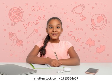 A Young Black African Girl With A Rapturous Look Is Writing Down Her Goals In A Notebook.  Doodle About Powers Of The Girl Are Drawn On The Pink Background. Happy Childhood Concept