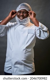 Young black African male chef putting on a hair net in studio wearing a chefs jacket black toque hat and beard net while smiling and laughing