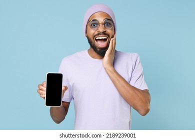 Young Black African Man 20s In Violet T-shirt Hat Glasses Show Mobile Cell Phone With Blank Screen Workspace Area Scream News With Hands Near Mouth Isolated On Pastel Blue Background Studio Portrait