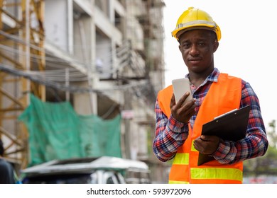 Young Black African Man Construction Worker Holding Clipboard And Mobile Phone At Building Site