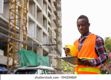 Young Black African Man Construction Worker Reading On Clipboard While Holding Hard Hat At Building Site