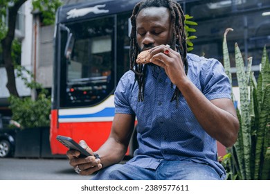young black african man with dreadlocks and beard, is sitting using mobile and checking phone and eating cookie outdoors. lifestyles concept, copy space - Powered by Shutterstock
