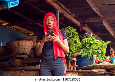 Young Black African Trader Using Phone In A Local Nigerian Market. Overexcited African Lady Reacting After Receiving Good News 