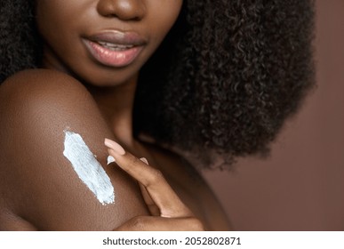 Young Black African woman applying moisturizing nutritive sunscreen lotion cream on body after shower on brown background. Skin body care products and spa bodycare concept. Closeup cropped image.