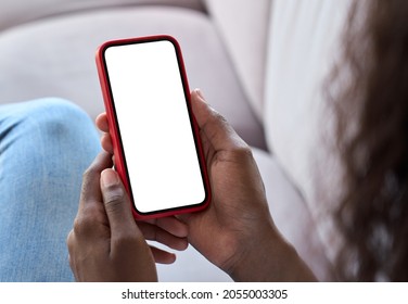 Young black African woman holding smartphone in hand with white black empty mockup screen template using cell phone relaxing on sofa at home. Over shoulder view. Mobile apps advertising concept.