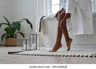 Young black African woman wearing bathrobe touching legs soft smooth skin applying lotion doing body care morning routine in bathroom. Spa, laser epilation hair removal, depilation. Close up