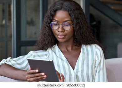 Young black African woman wearing glasses holding digital tablet sitting on couch at home learning online, watching videos, surfing internet, doing e shopping using technology device relaxing on sofa. - Shutterstock ID 2055003332