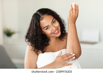 Young Black Bodypositive Woman In Towel Applying Soothing Cream At Elbow, Moisturizing Dry Skin At Home, Copy Space. Happy Lady Pampering Herself At Bathroom