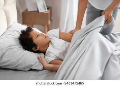 A young black boy lies in bed with a tousled head of hair, gazing up at an unrecognizable adult standing beside the bed, who is gently pulling back the duvet - Powered by Shutterstock