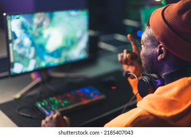 Young Black Boy In Orange Hoodie Sitting In Front Of Screen Giving Instructions To Teammates Playing An Online Game. Gaming Computer Setup. High Quality Photo