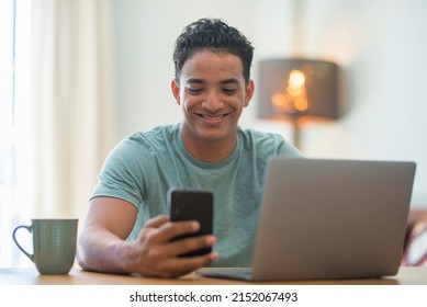 Young Black Boy Using Phone And Computer Connection At Home. Happy Man Reading Notification On Mobile Cellular While Work On Laptop In Smart Working. Modern Z Generation People Using Web