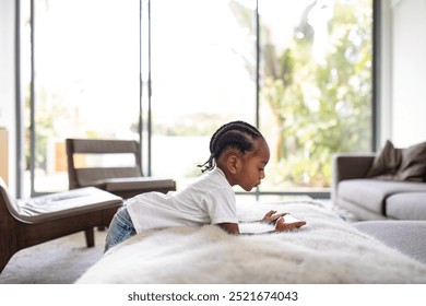 Young Black boy in a white shirt, playing on a phone. The boy is focused on the tablet, lying on a couch. Bright, modern living room with large windows. African American boy using phone in living room - Powered by Shutterstock