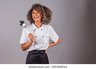 Joven brasileña negra, cocinera, maestra, con uniforme de restaurante. sostener una cuchara grande para preparar salsas.