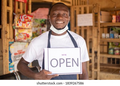 Young Black Business Owner Wearing A Face Mask And Holding An Open Sign In Front Of His Shop And Smiling