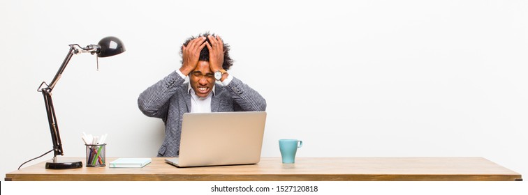 young black businessman feeling stressed and anxious, depressed and frustrated with a headache, raising both hands to head on a desk
