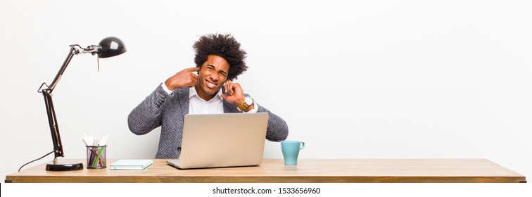 Young Black Businessman Looking Angry, Stressed And Annoyed, Covering Both Ears To A Deafening Noise, Sound Or Loud Music On A Desk