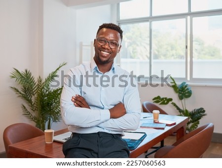 A young Black businessman poses with arms folded at conference table in office
