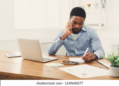 Young black businessman talking on mobile phone and making notes while working on laptop in modern white office, copy space - Shutterstock ID 1149640577