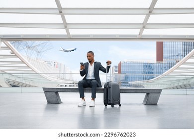Young Black Businessman Using Smartphone While Waiting Flight At Airport Hall, African American Male In Suit Texting On Mobile Phone While Sitting On Bench At Terminal, Plane Departure On Background