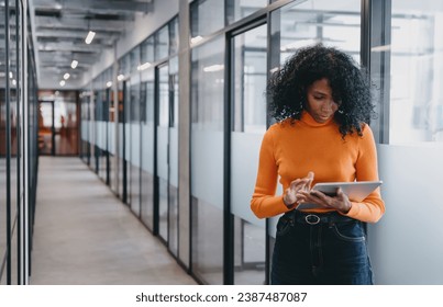 Young Black businesswoman using tablet in modern office corridor, focused and serious. Wearing casual work attire, she embodies professionalism, technology and mobility in modern business. - Powered by Shutterstock