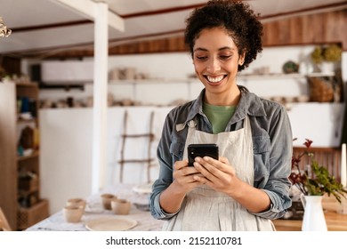 Young black ceramist woman wearing apron using cellphone while working in her studio - Powered by Shutterstock