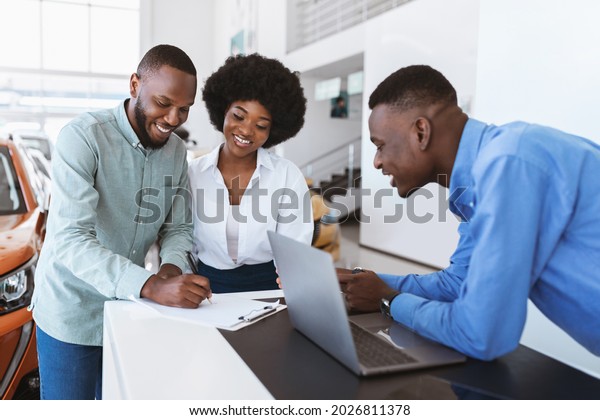 Young black couple\
buying new auto, signing car purchase agreement at dealership.\
Happy African American customers making deal with salesman at\
automotive showroom store